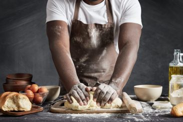 Cropped shot of black male wears apron, kneads dough as prepares delicious rolls, going to make surprise for family and treat with tasty pastry, isolated over black background. Baking concept Cropped shot of black male wears apron, kneads dough as prepares delicious rolls, going to make surprise for family and treat with tasty pastry, isolated over black background. Baking concept